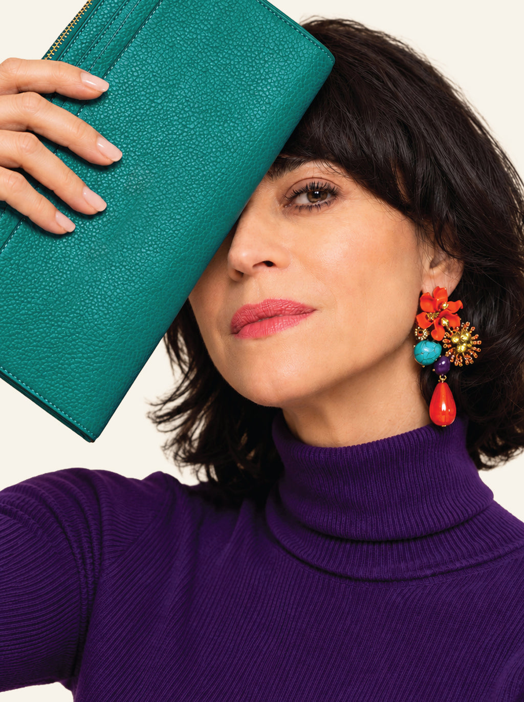 A woman with dark hair wearing a purple turtleneck sweater and a colorful statement earring, holding a teal textured leather clutch in front of her face against a light background.