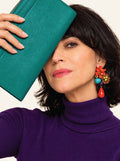 A woman with dark hair wearing a purple turtleneck sweater and a colorful statement earring, holding a teal textured leather clutch in front of her face against a light background.
