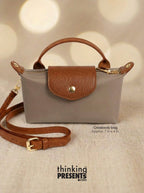 Taupe crossbody with brown handle and strap on a beige background, featuring 'thinking PRESENTS'.com' branding.