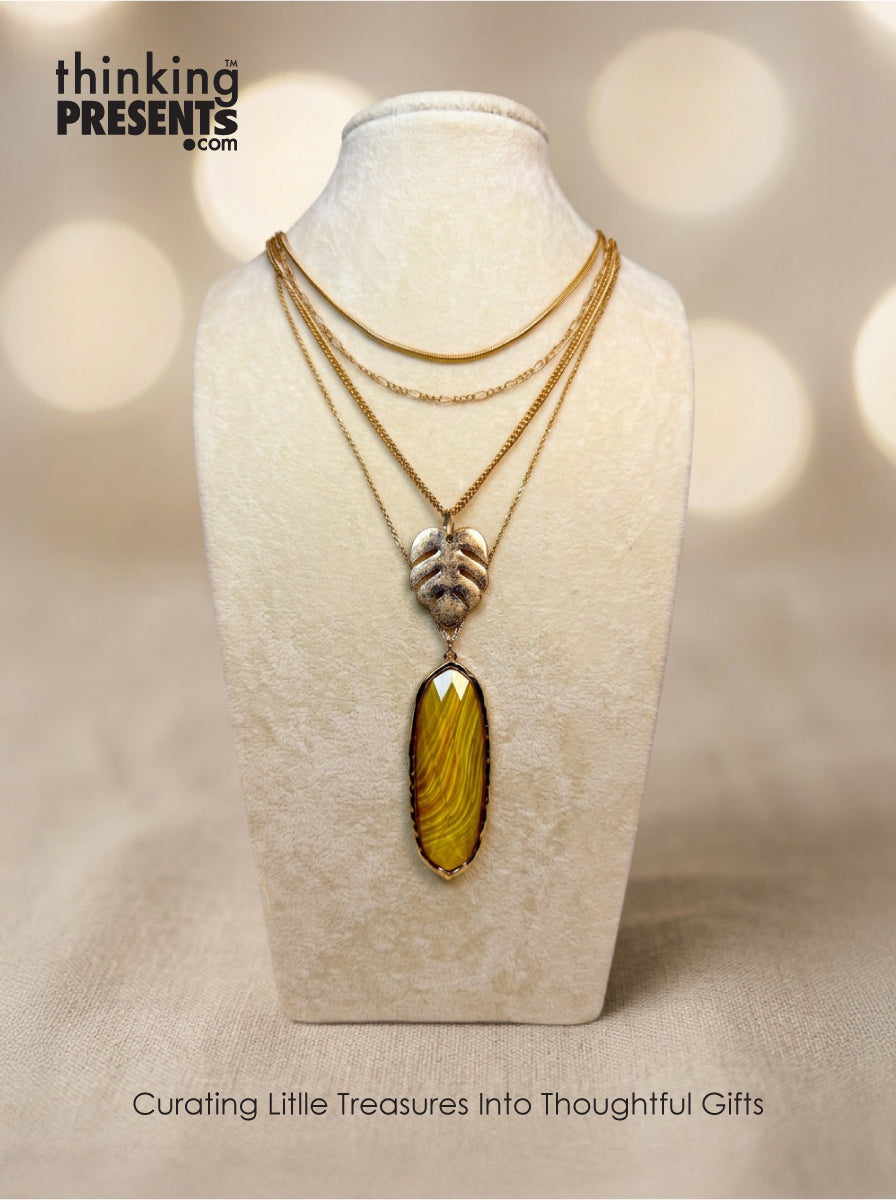 Layered gold-tone necklace with textured chains, leaf motif detail, and elongated amber-toned pendant, styled on a neutral display, warm and nature-inspired curated gift aesthetic